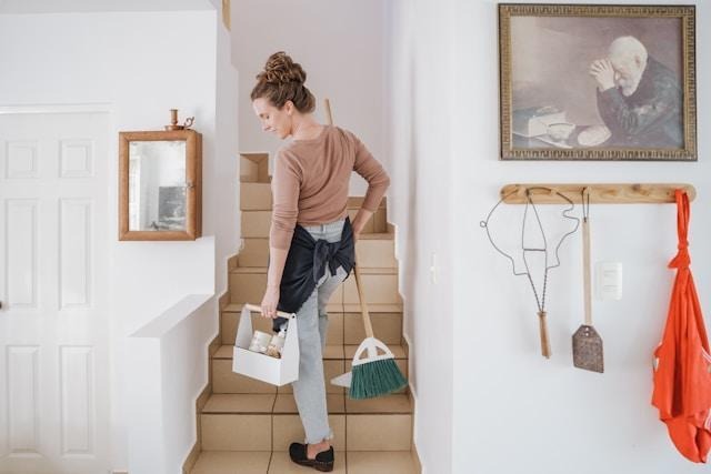 A woman walking up the stairs with a broom and cleaning supplies.